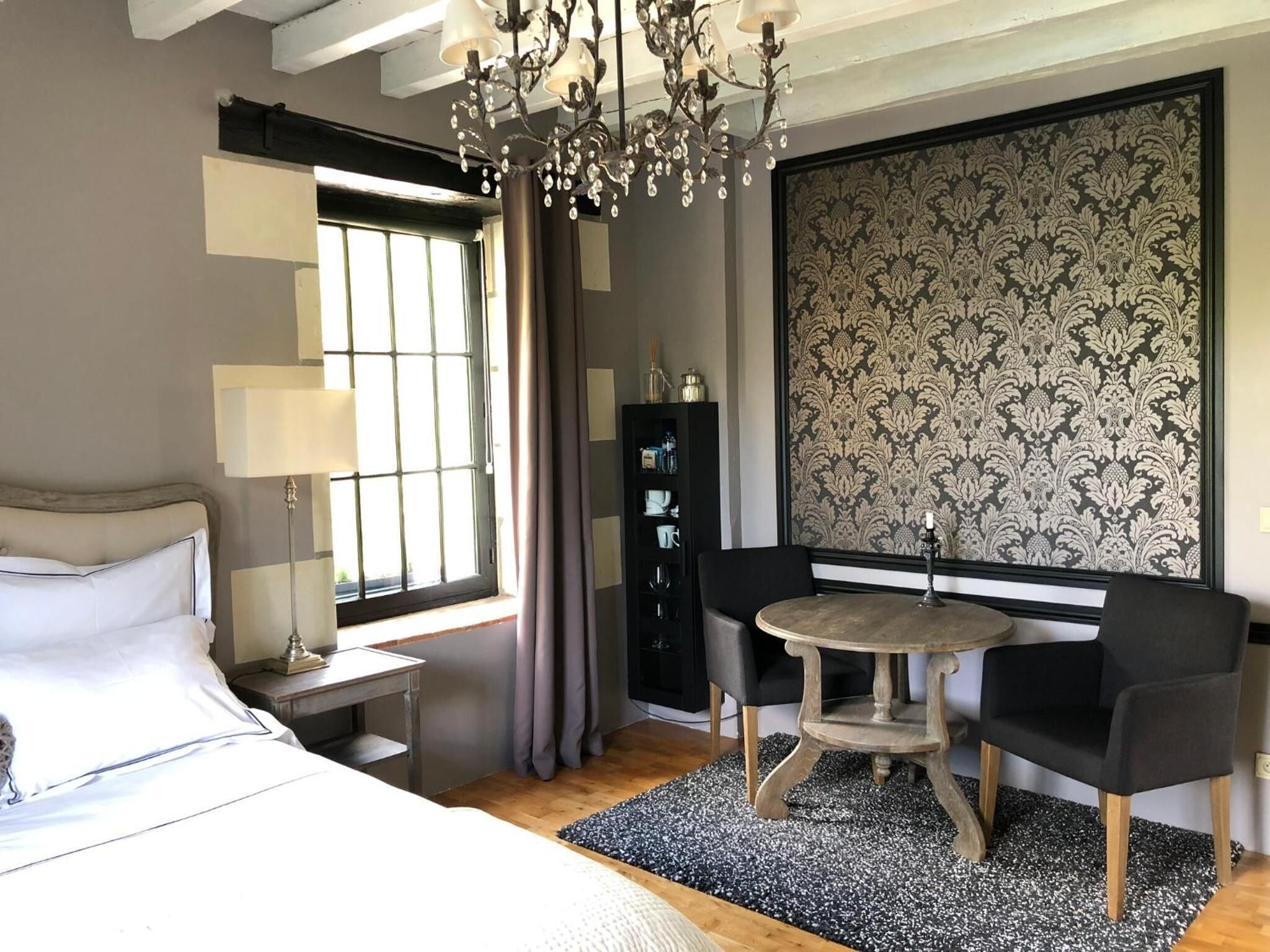 Una habitación de hotel moderna con una cama, una mesa redonda con dos sillas, una araña, un ventanal con cortinas y un panel decorativo en la pared.