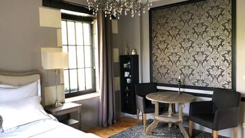 Una habitación de hotel moderna con una cama, una mesa redonda con dos sillas, una araña, un ventanal con cortinas y un panel decorativo en la pared.