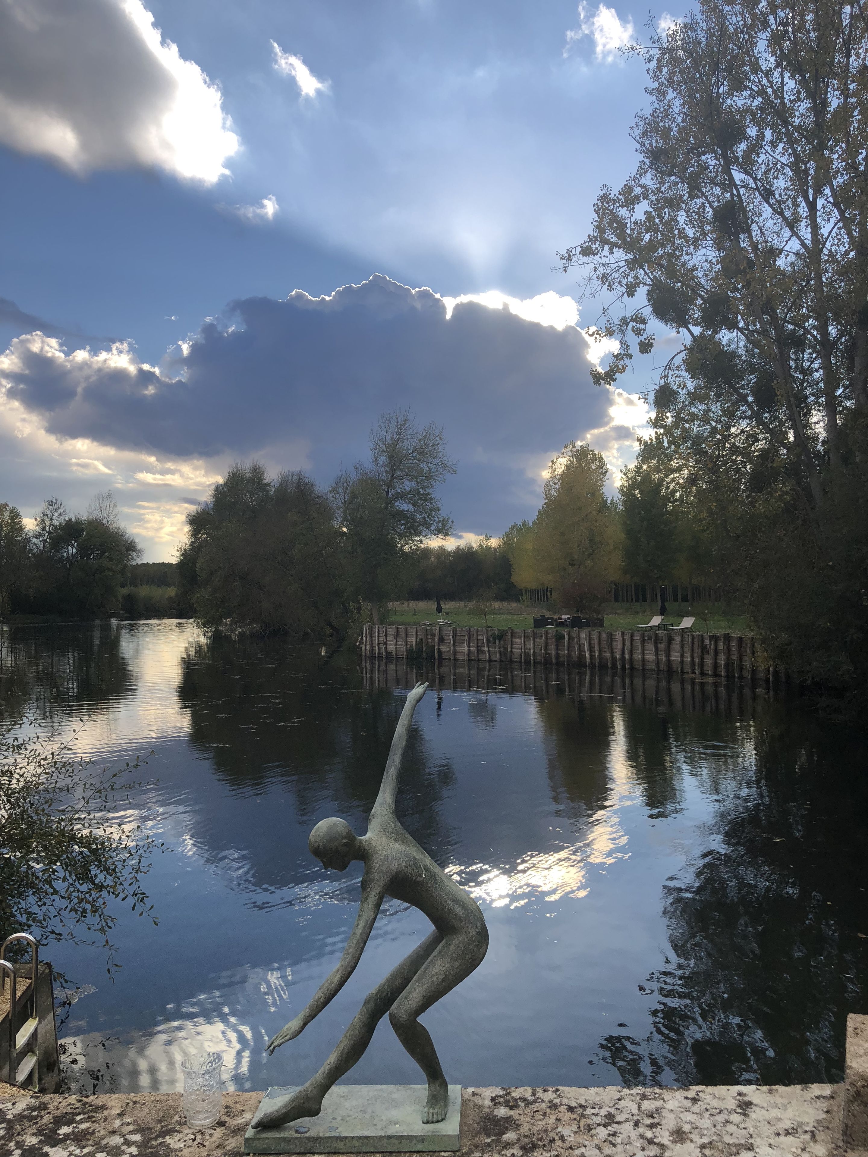 Una escultura de figura humana junto a un río tranquilo, rodeada de árboles y un cielo nublado.