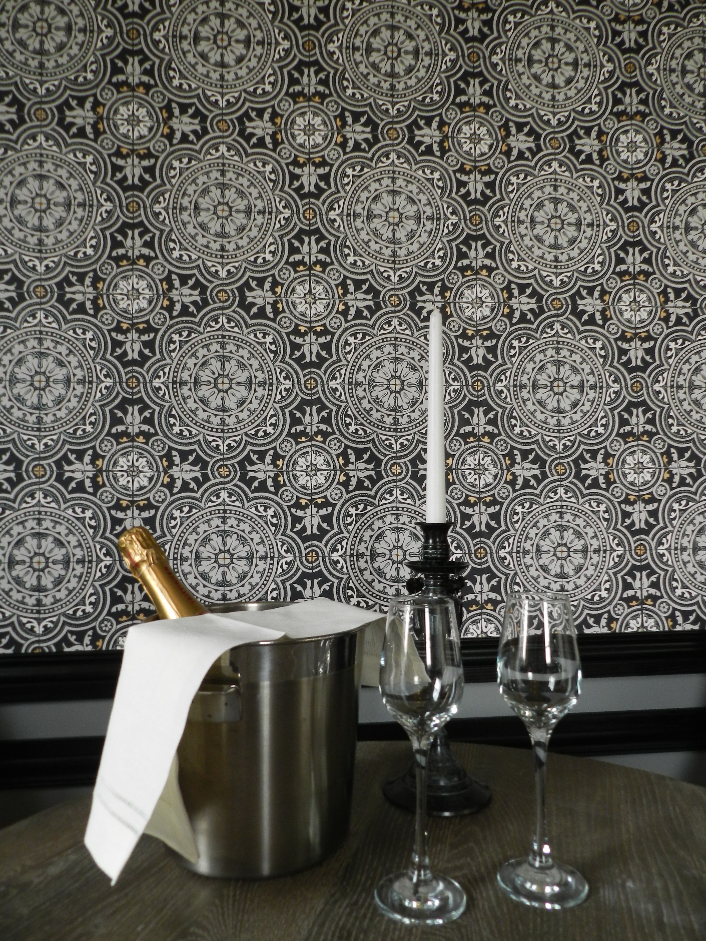 A table with a bottle of champagne, a candle, and two wine glasses against a patterned wallpaper background.