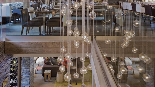 Un vaste espace à manger doté de hauts plafonds et de poutres en bois, agrémenté de luminaires suspendus et d’une balustrade en verre donnant sur un niveau inférieur.