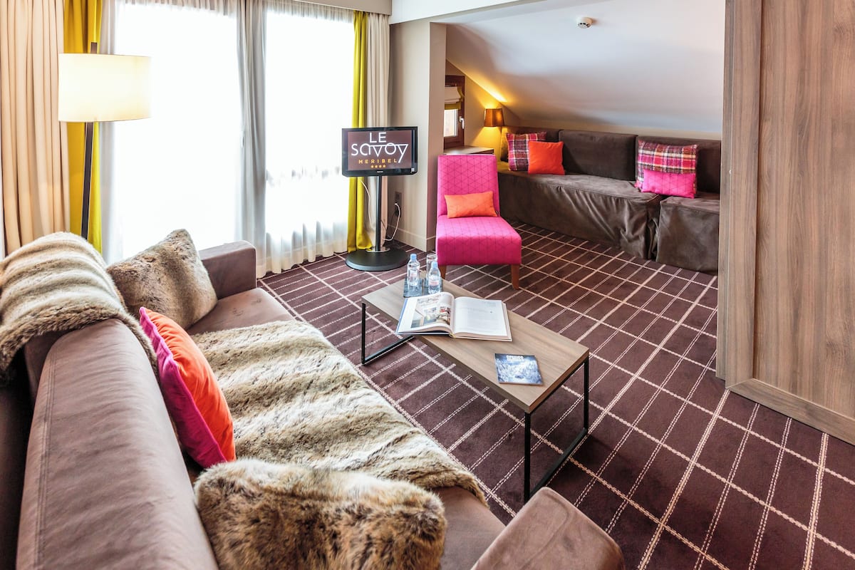 Hotelkamer met een bruine bank, een roze stoel, een houten salontafel met een tijdschrift en een televisiescherm waarop 'LE SAVOY' staat.