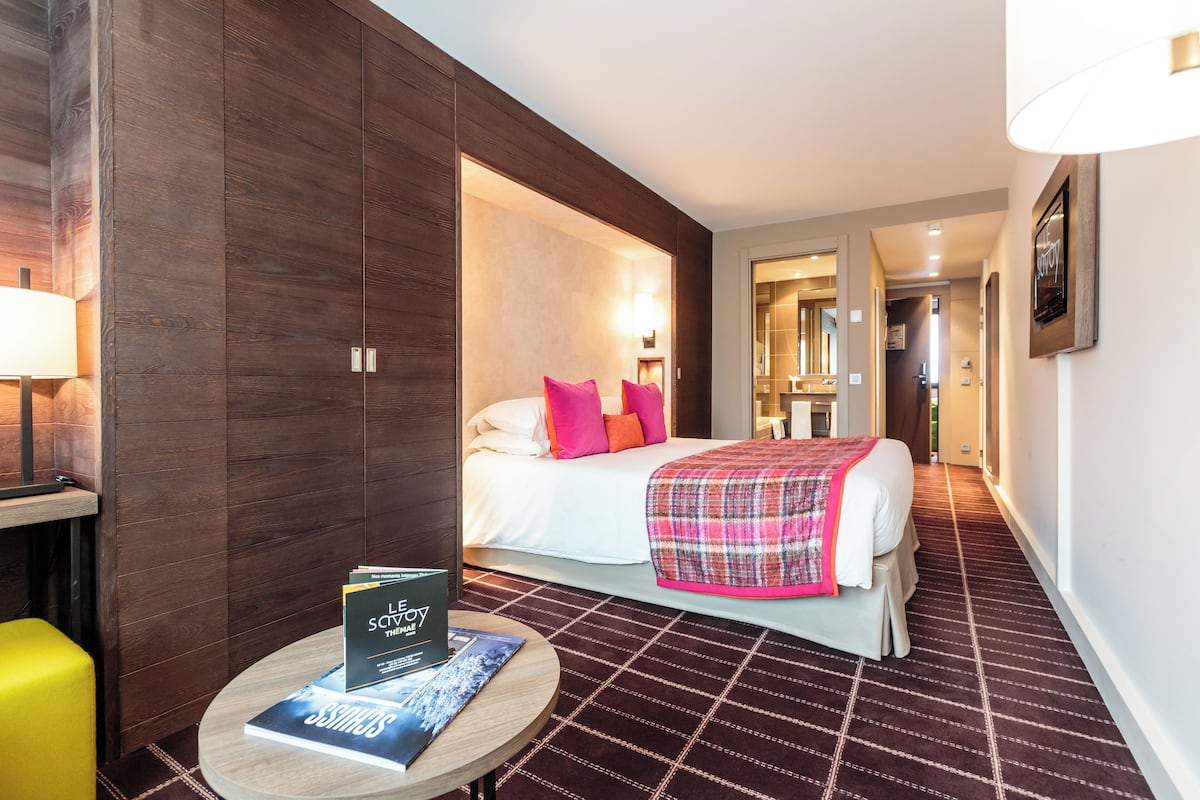 Een hotelkamer met een bed, een nachtkastje met een lamp, een ronde tafel met een boek en een houten garderobe.