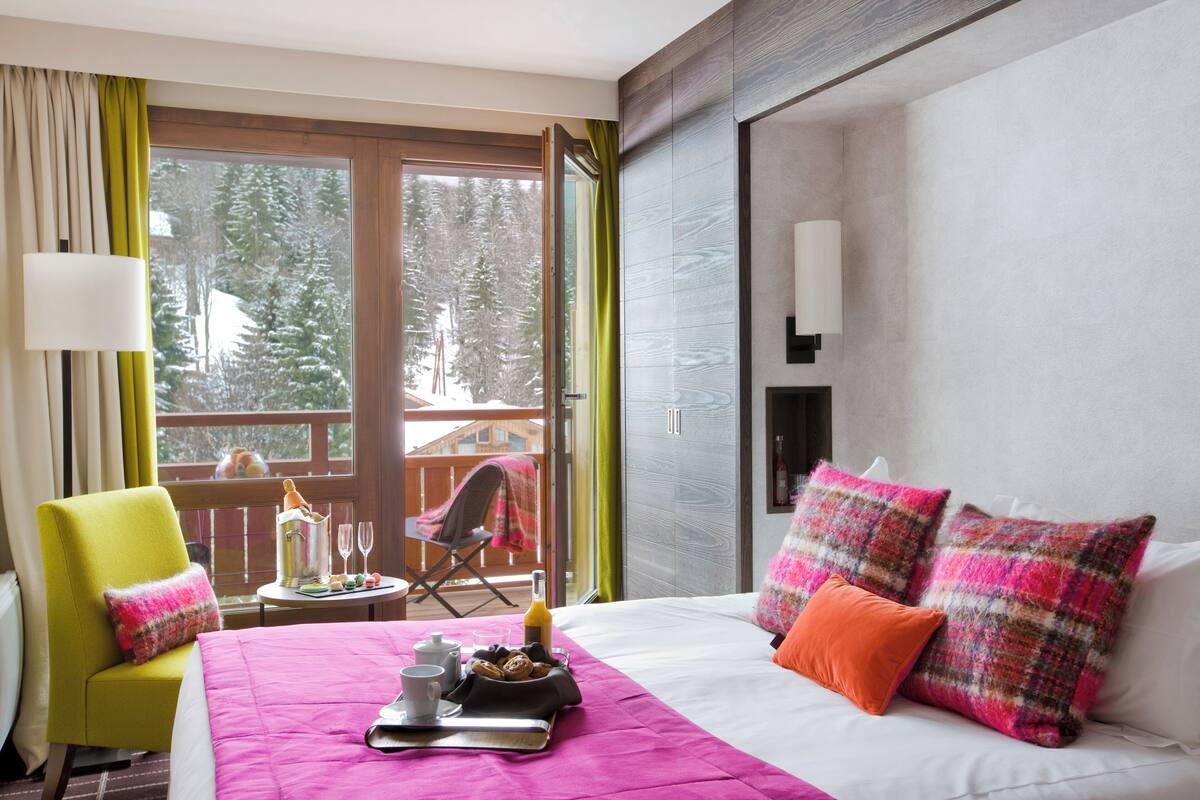 Een hotelkamer met een bed, een stoel, een tafel met ontbijtartikelen en uitzicht op bomen door een groot raam.