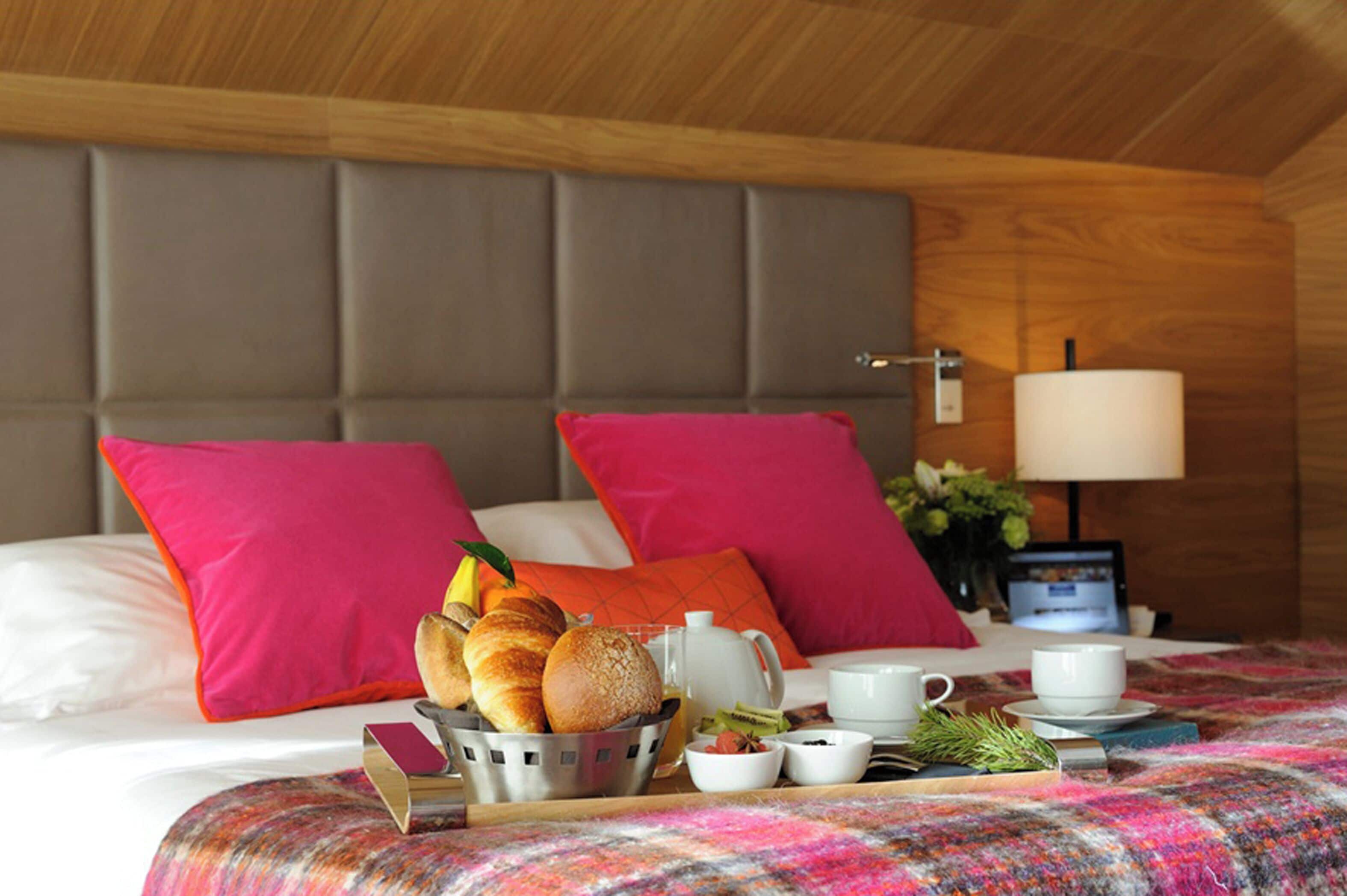 Hotelkamer met een bed, kussens, een nachtkastje met een lamp en een ontbijttafel met brood, fruit en kopjes.
