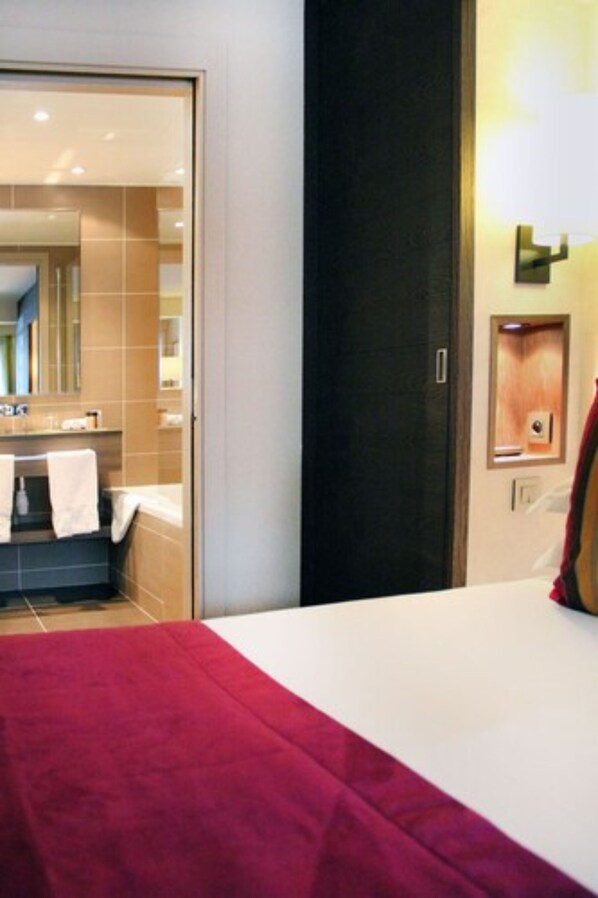 A hotel room with a bed, a bathroom with a mirror, and a wall-mounted light fixture. - Hôtel Le Savoy Méribel (Les Allues)