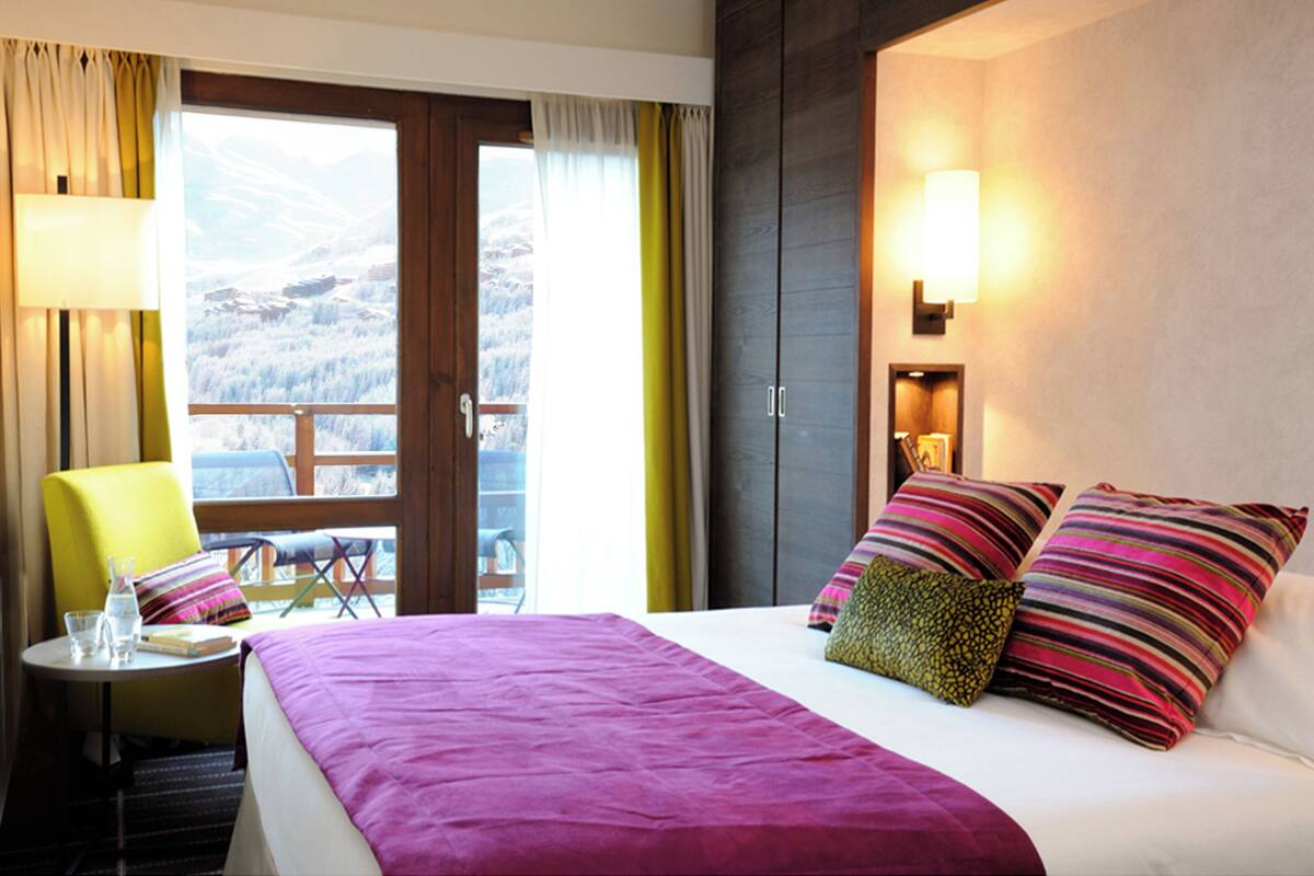 Hotelkamer met een bed, een stoel, een tafel met een glas water en uitzicht op bergen door het raam.