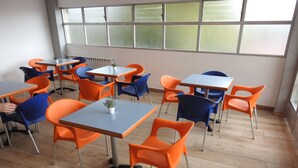 Une salle de classe avec des chaises et des tables orange et bleues, de grandes fenêtres et un radiateur.