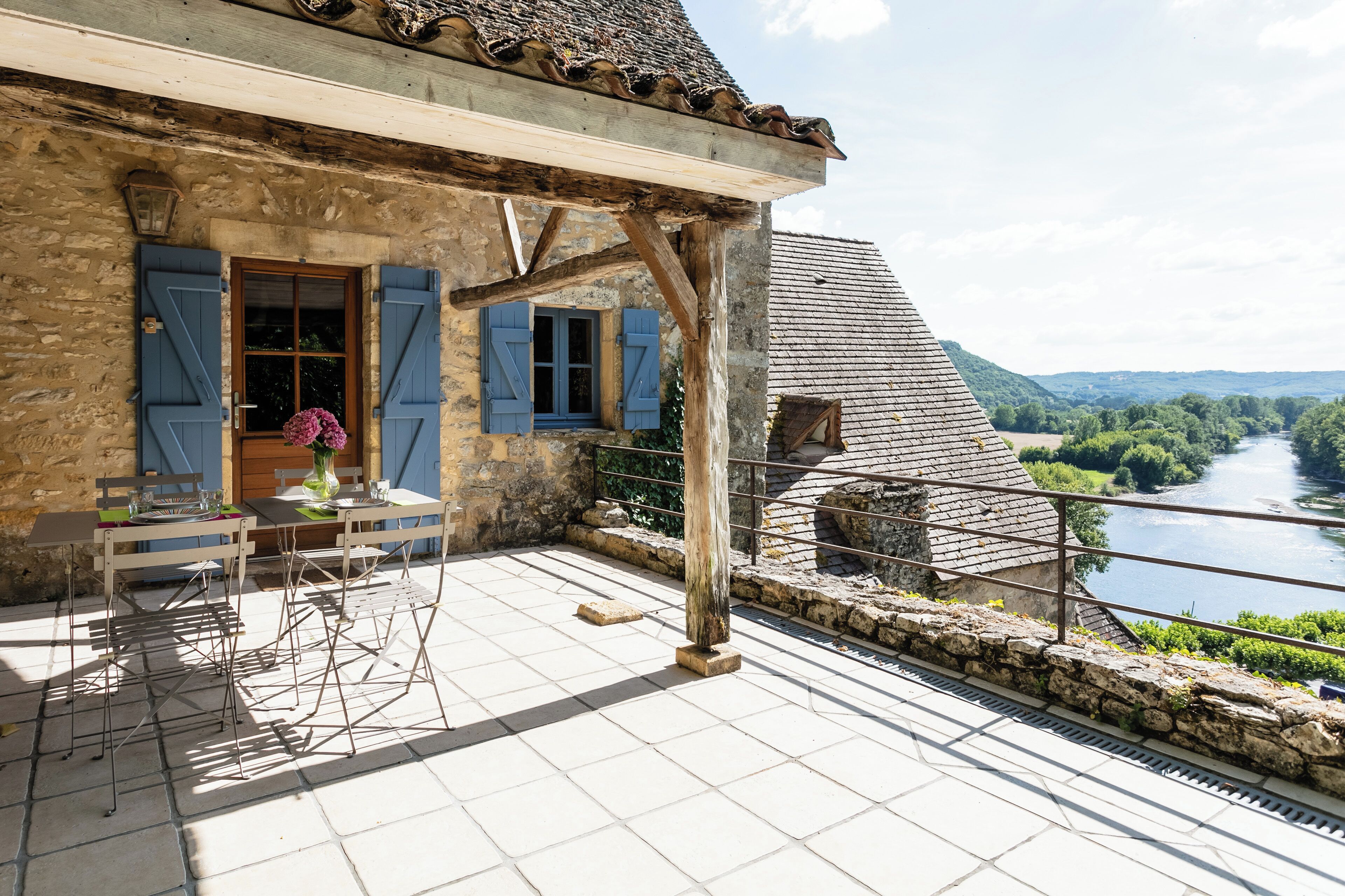 A stone building with blue shutters, a balcony with a table and chairs, and a view of a river and hills.