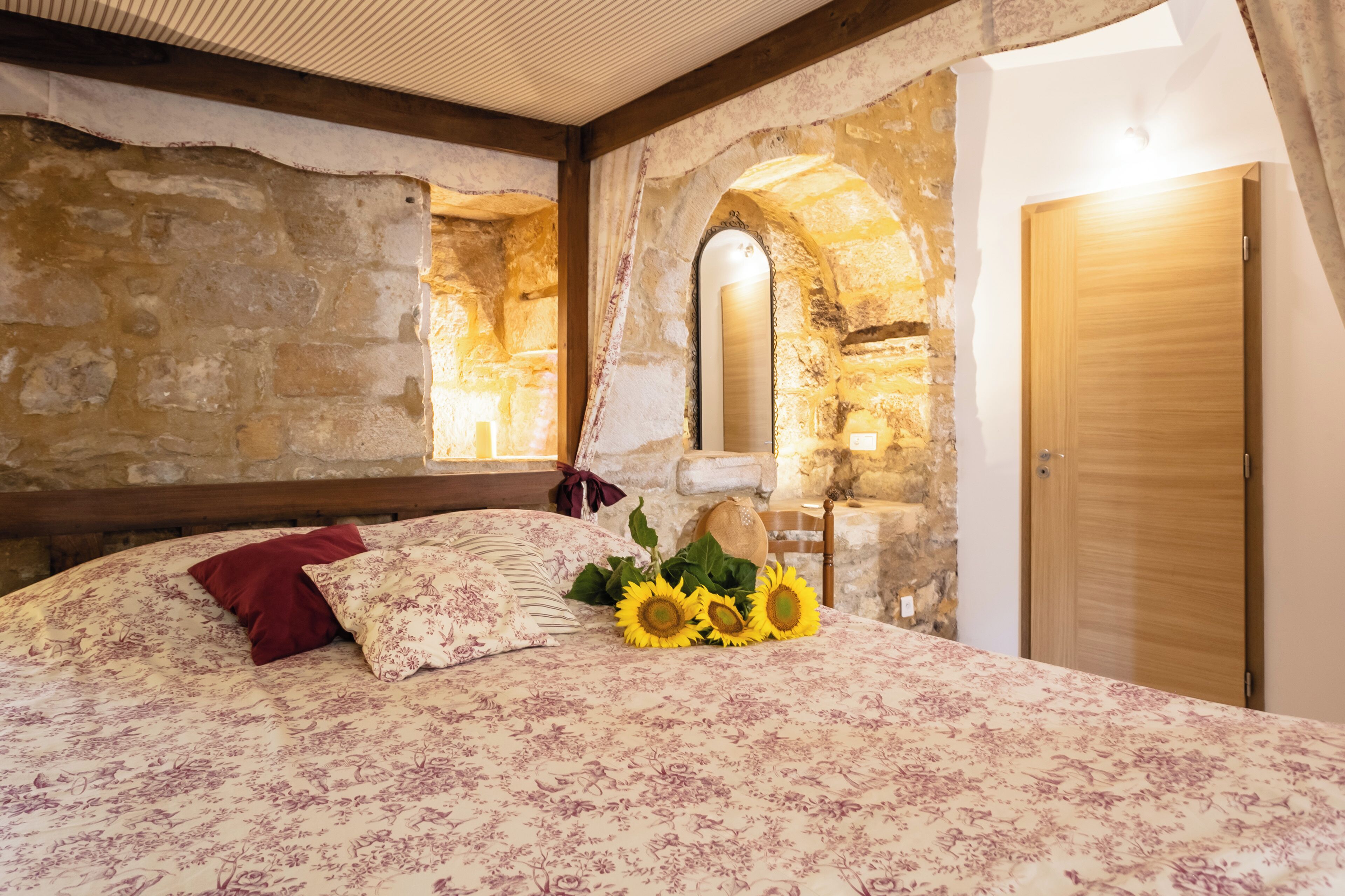 A bedroom with a stone wall, a wooden door, and a bed with floral bedding and sunflowers.