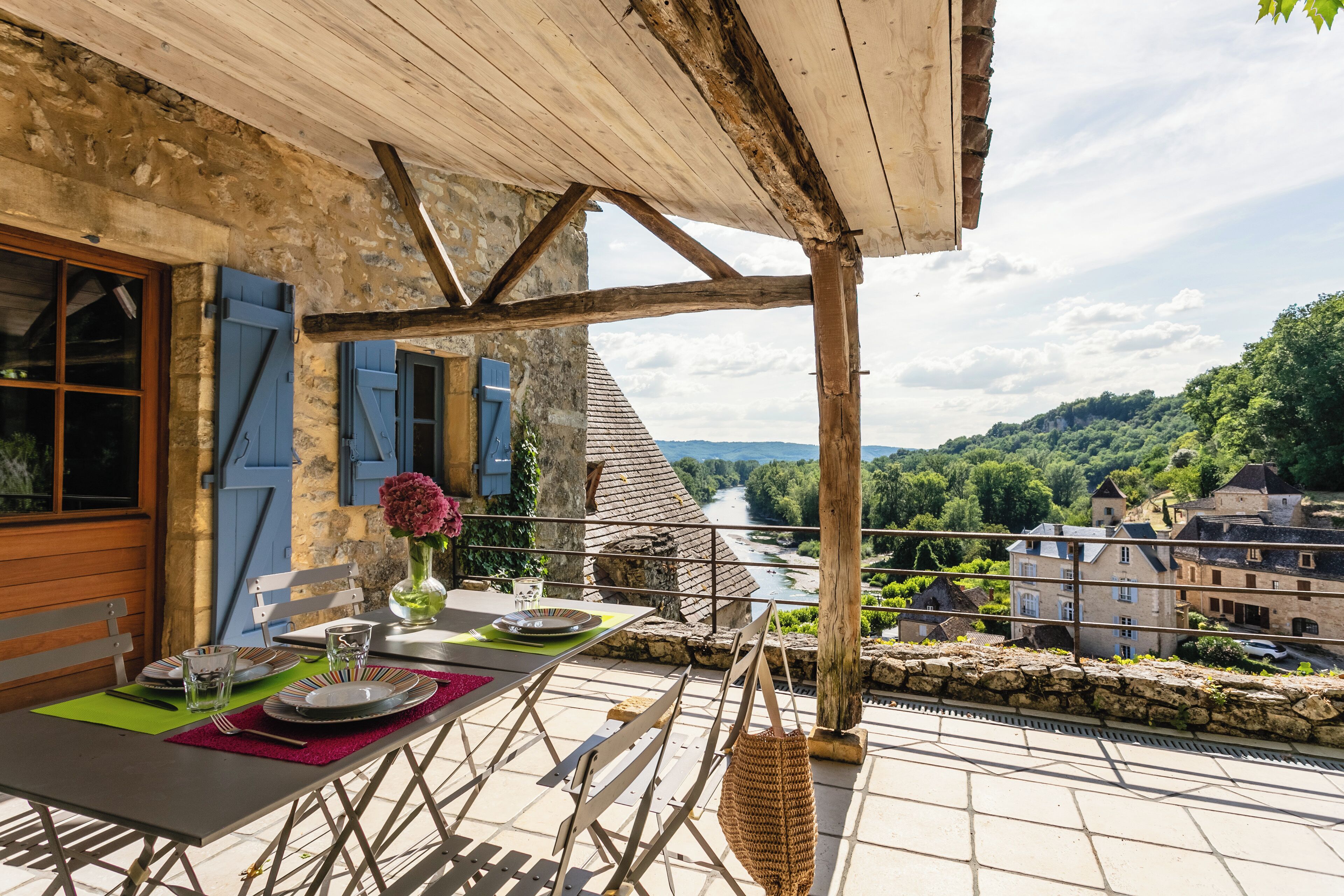 Une terrasse avec une table dressée pour le dîner, offrant une vue sur une rivière et des bâtiments.