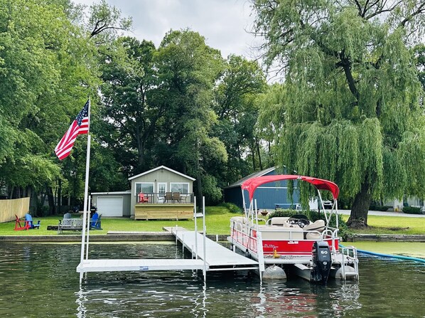 A docked boat with a red canopy, a small house with a porch, and an American flag. - Lakehouse with Pontoon, Deck, Firepit, Paddle Boards, Grill & more INCLUDED!!  (Valparaiso)