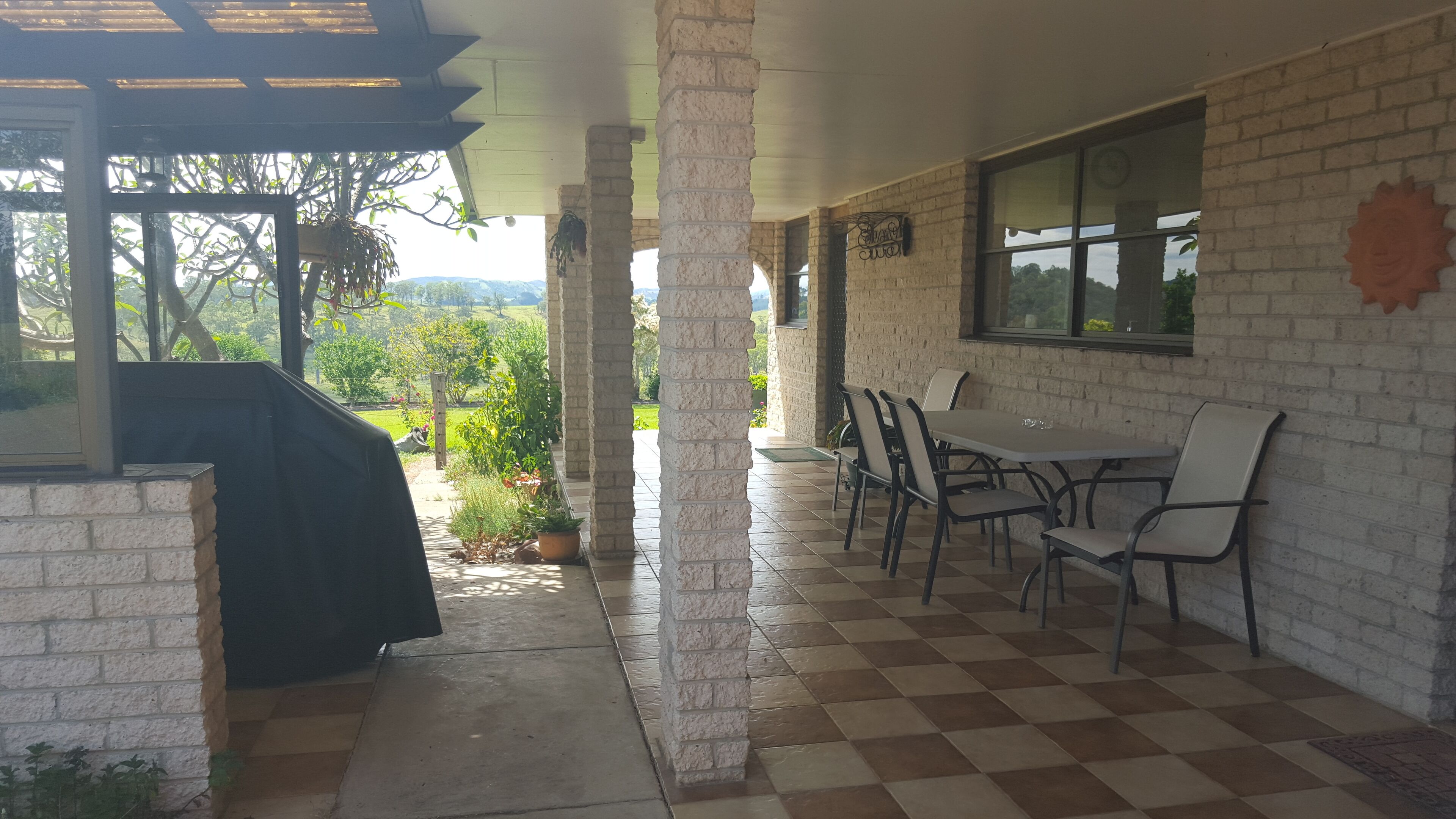 Un patio couvert avec une table et des chaises, un barbecue, et une vue sur un jardin et des collines en arrière-plan.