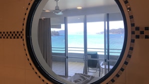 A circular mirror reflecting a balcony with a view of the sea, a ceiling fan, and a chair.