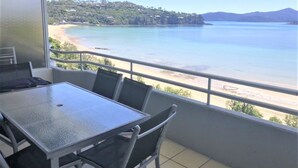 A balcony with a glass table and chairs overlooking a beach and a mountainous landscape.