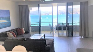 A modern living room with a large window overlooking a scenic ocean view, a dining area with a table and chairs, and a sofa with cushions.