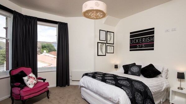 A bedroom with a bed, a chair, a window with curtains, a wall with framed pictures, and a sign with the name 'LICKERISH 373'. - Red Door Penthouse in Battery Point (Battery Point)