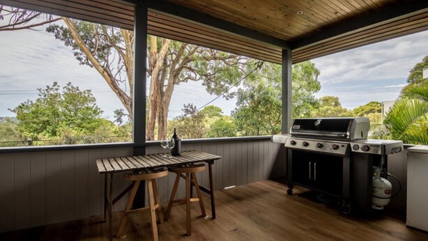 A covered outdoor area with a wooden table and two stools, a barbecue grill, and a gas cylinder. - Woodlands at Beachview by HolidayCo. (MACMASTERS BEACH)