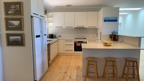 A modern kitchen with white cabinets, a built-in oven, a coffee machine, and a fruit bowl on the counter. There are three wooden stools and a small island with stools. - Woodlands at Beachview by HolidayCo. (MACMASTERS BEACH)