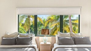 Un dormitorio con dos camas, una mesita de noche, una lámpara y vistas a palmeras a través de amplias ventanas.