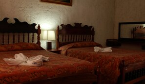 Ein Hotelzimmer mit zwei Betten, jeweils mit Holz Kopfteil und einem gemusterten Bettüberwurf. Ein Nachttisch mit Lampe und ein Spiegel an der Wand sind vorhanden.