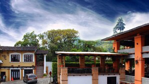 A courtyard with a covered seating area, a car parked in front, and a building with a red facade and a balcony. - Hotel Alcazar De Doña Victoria (Cobán)