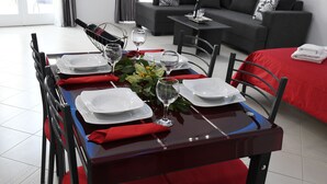 A dining table set for four with white plates, red napkins, and wine glasses, in a modern room with a grey sofa and a red bedspread.