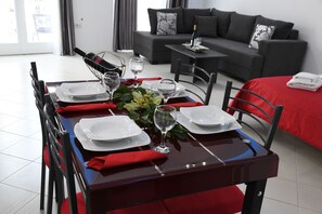 Une table à manger dressée pour quatre personnes, avec de l’assiette blanche, des serviettes rouges et des verres à vin, dans une pièce moderne avec un canapé gris et une couverture de lit rouge.