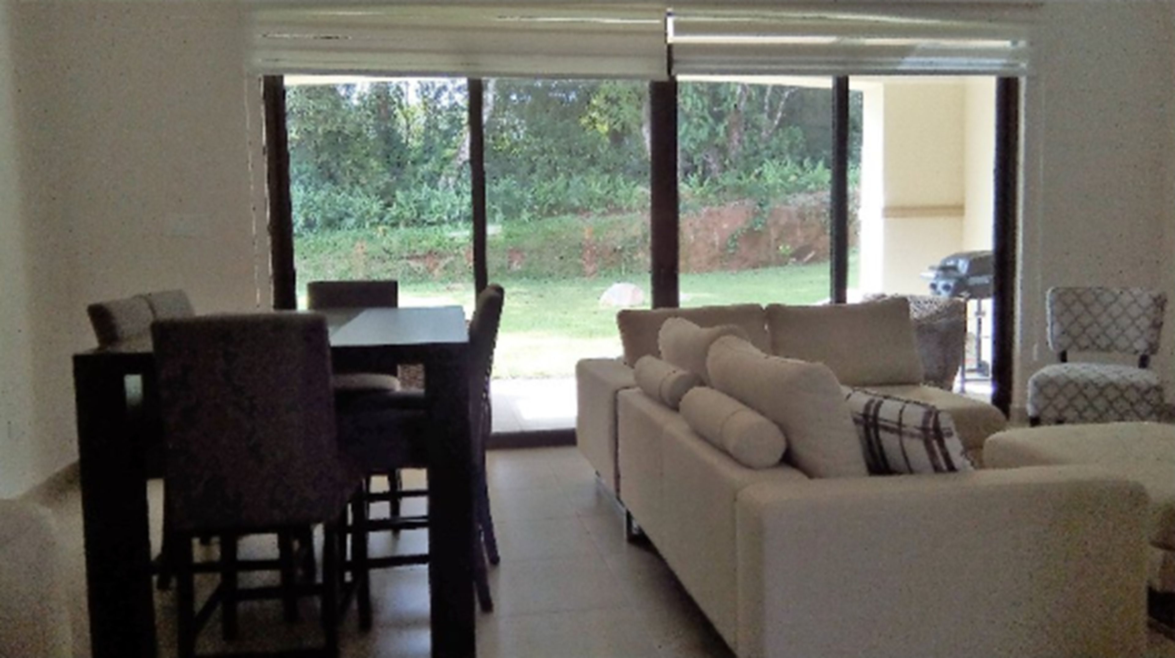 Una sala de estar con un sofá, una mesa de comedor con sillas y vistas a un jardín a través de puertas de vidrio grandes.