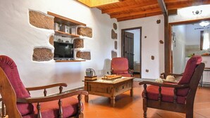 A cozy living room with stone walls, wooden furniture, a television, and a kitchen visible through an open door. - GC0242 Holiday cottage with private pool in Santa Lucía (Santa Lucía de Tirajana)