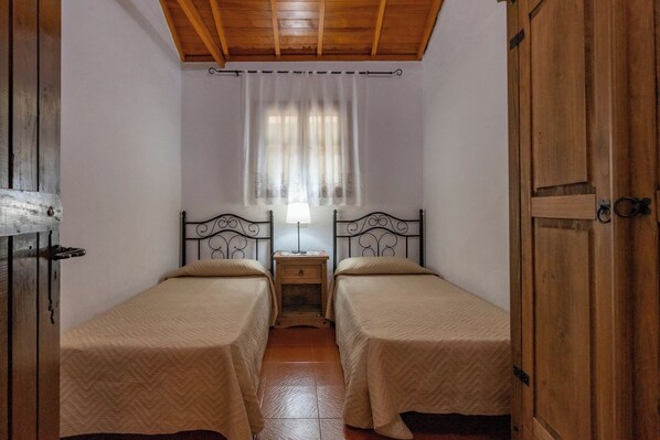 A room with two single beds, wooden ceiling, white walls, a window with sheer curtains, and a nightstand with a lamp.