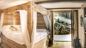 A wooden four-poster bed with white linens, a rustic wooden floor, and a view of a rocky outdoor area through an open doorway. - Charming lux chalet, views, comfort, escape, Samoens Valley, peaceful log fire (Mieussy)