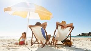 Three people relaxing under a beach umbrella on a sandy beach.