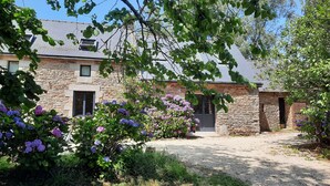 Un bâtiment en pierre avec une allée de gravier, entouré d’une végétation luxuriante et de plantes en fleurs.