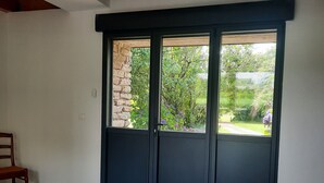 Une chambre à coucher avec un grand lit, une chaise en bois et une vue sur un jardin, visible à travers une porte de couleur sombre.