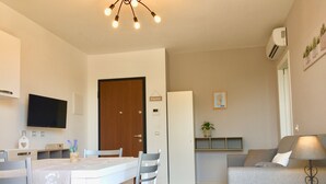 Ein modernes Wohnzimmer mit grauem Sofa, einem Esstisch mit Stühlen, einem an der Wand montierten Fernseher und einer Deckenleuchte.