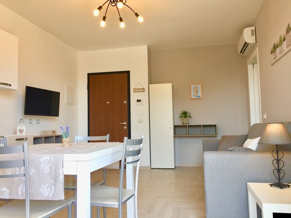 Un salon moderne avec un canapé gris, une table à manger avec des chaises, un téléviseur fixé au mur et un luminaire suspendu au plafond.
