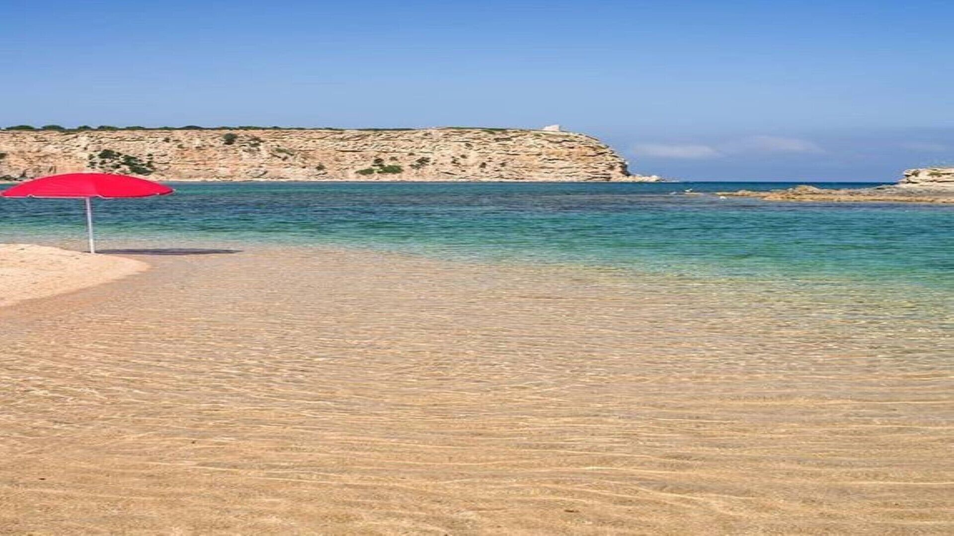 A beach with clear shallow waters, a red umbrella, and a rocky cliff in the background.