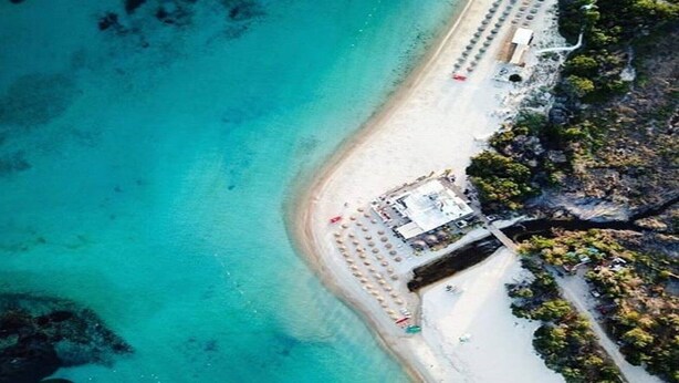 Ein Luftbild zeigt einen Strand mit weißem Sand, klares türkisfarbenes Wasser und ein kleines Gebäude mit flachem Dach am Ufer.