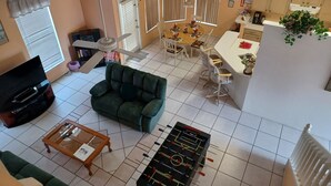Un salon comprenant un canapé, une table basse, un téléviseur et un baby-foot. On y trouve également un coin repas avec une table et des chaises, ainsi qu’une cuisine équipée de placards et d’un évier.