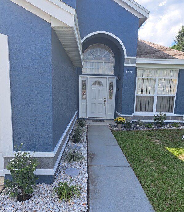 A two-story house with a blue exterior, a white front door with a glass panel, and a small garden area with gravel and plants. - 4BR Private Pool Home Near Disney – Family Friendly Sleeps 8 Screened Pool (Kissimmee)