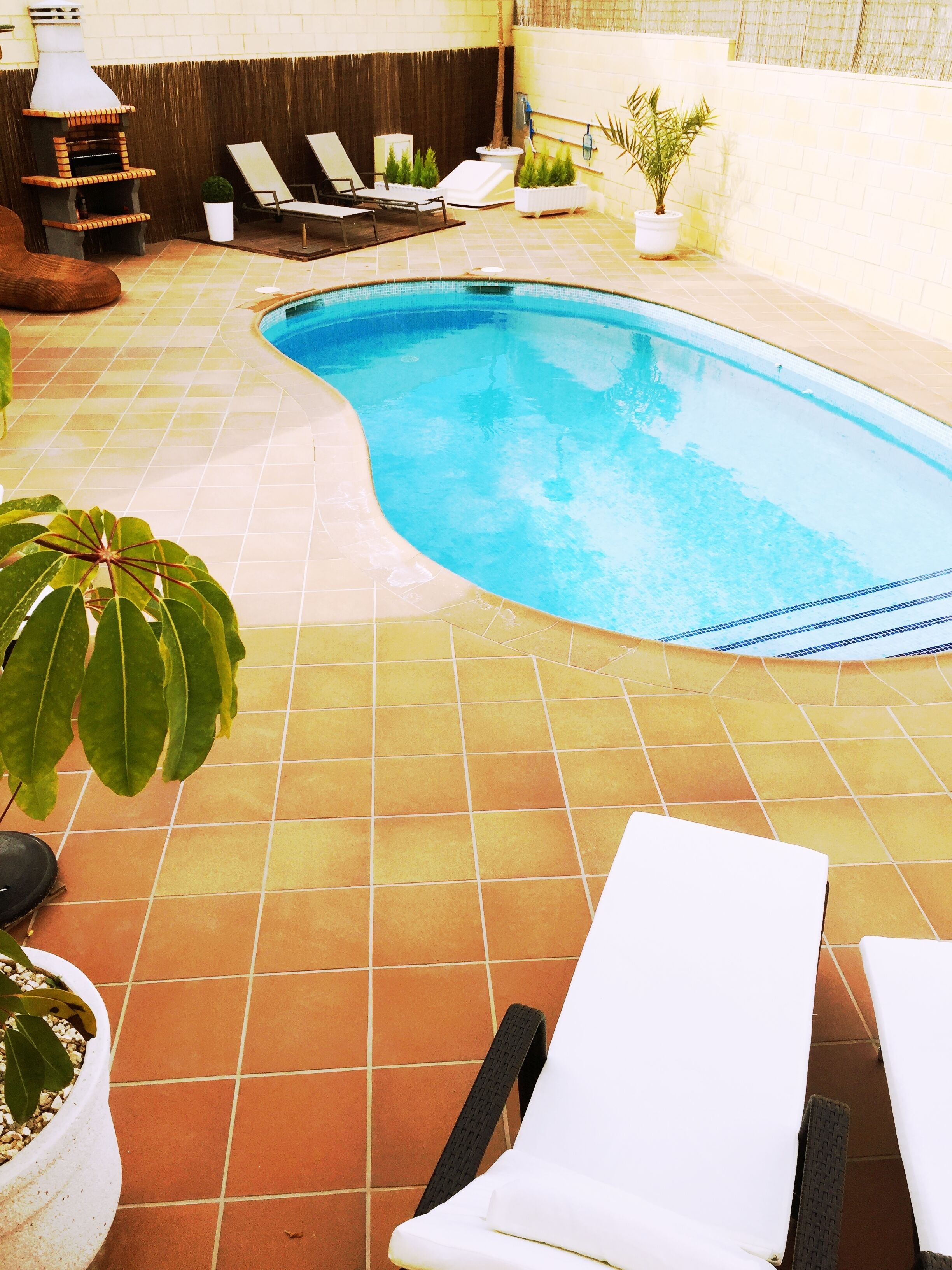 Une piscine avec un sol carrelé, des chaises longues et des plantes en pot.