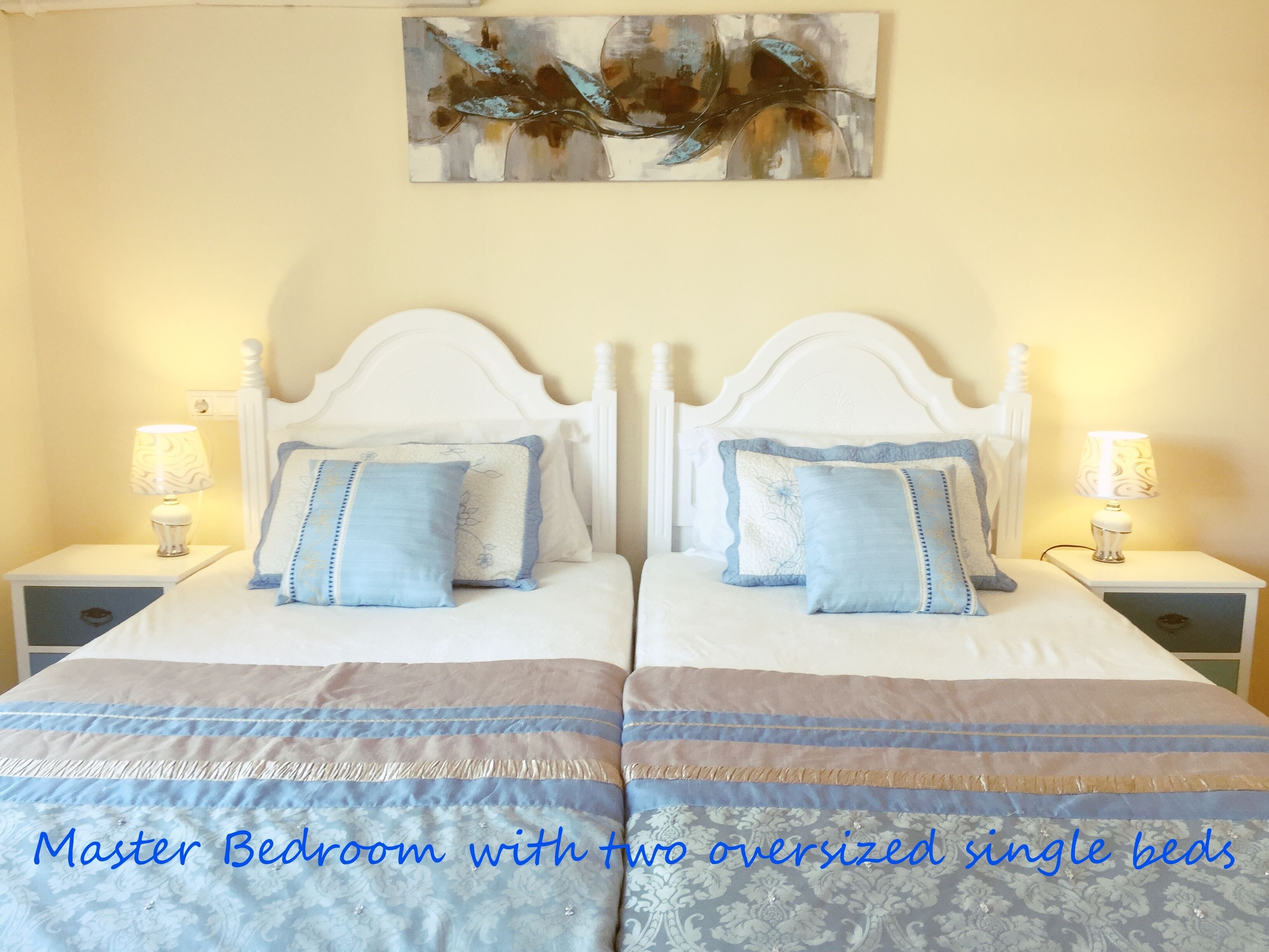 Ein Schlafzimmer mit zwei besonders großen Einzelbetten, jeweils mit weißem Kopfteil, blauer und weißer Bettwäsche sowie je einer Nachttischlampe.