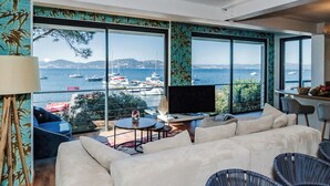 Ein modernes Wohnzimmer mit einem großen Fenster, das Blick auf Boote und eine entfernte Küstenlinie bietet.