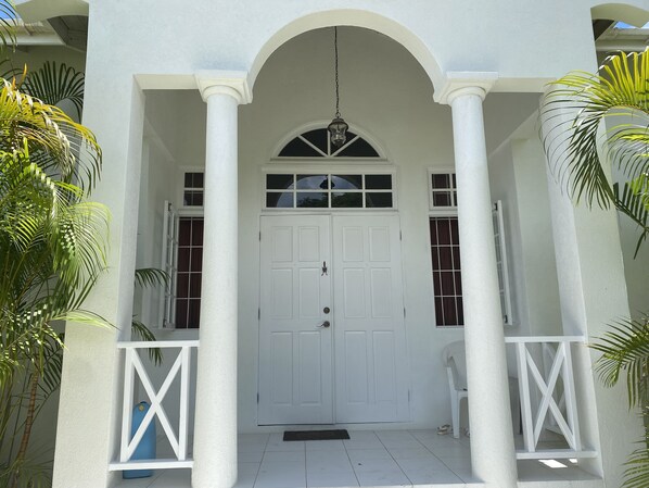 A white building with a central door, two windows on either side, and a small balcony with a chair. - Villa, 4 beds, 3 bathrooms, 1 minute to beach by car or 10 minute stroll (Westmoreland)