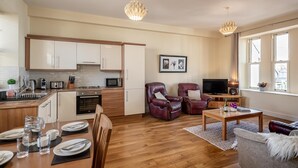 A modern kitchen with white cabinets, a dining area with a wooden table set for four, and a living area with leather chairs and a TV. - A 2 bed apartment that sleeps 4 guests  in 2 bedrooms (kenmare)