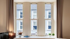 A room with a large window, a television on a stand, a small table with a lamp and decorative items, and a fluffy chair. - A 2 bed apartment that sleeps 4 guests  in 2 bedrooms (kenmare)