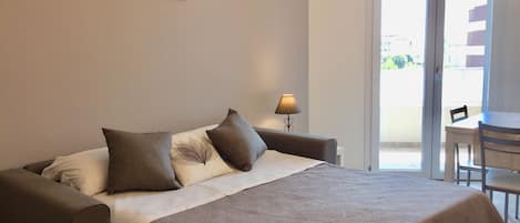 A bedroom with a bed, a nightstand, a lamp, a dining table, and a view of the outside.