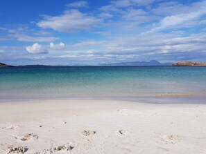 Una playa con agua cristalina, arena blanca y montañas a lo lejos.
