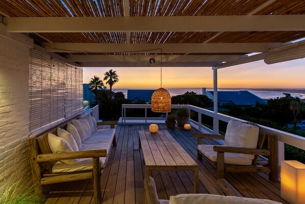 Eine Holzterrasse mit einer Couch und Stühlen, einer Hängeleuchte und Blick auf den Ozean bei Sonnenuntergang.
